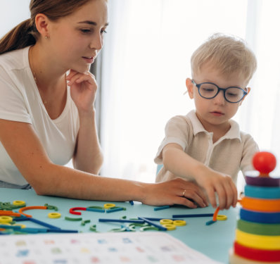 A woman and a young boy with glasses engage in a playful learning activity at a table, stacking colorful rings.