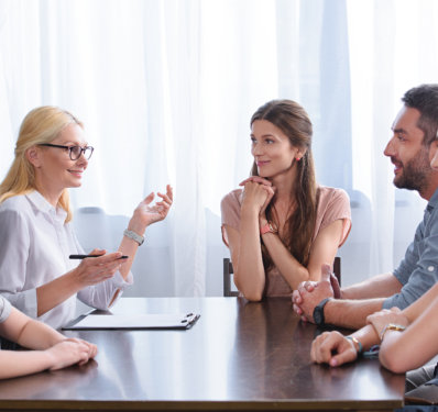 A woman with glasses discusses around a table, with a person gesturing and explaining while others listen attentively in a bright, airy room.