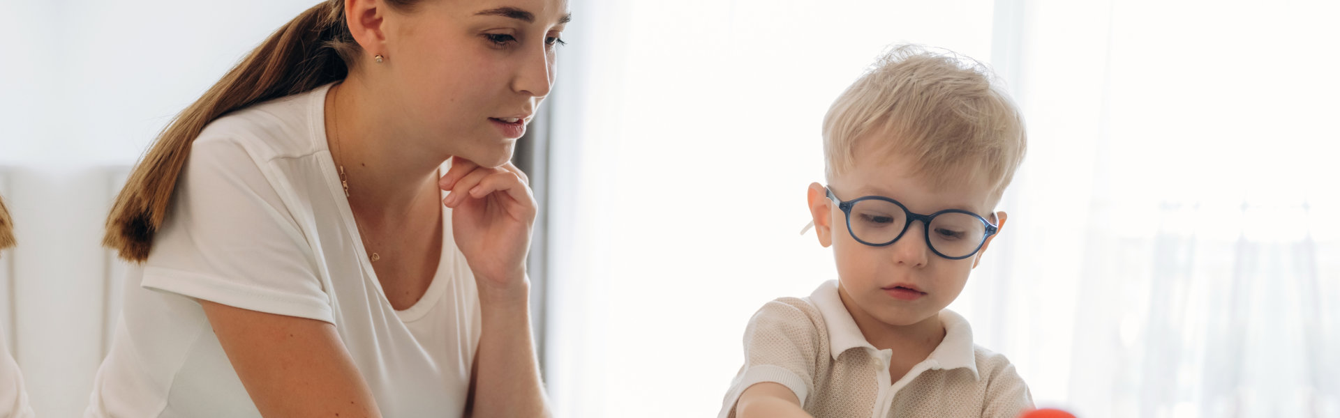 A woman and a young boy with glasses engage in a playful learning activity at a table, stacking colorful rings.